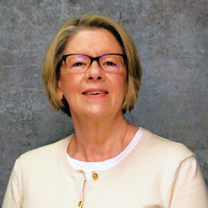 Friendly woman with medium length blonde hair smiling against a textured gray backdrop in a tan sweater.