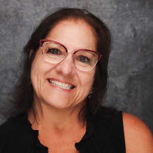 Friendly woman with medium length dark hair smiling warmly against a textured gray backdrop in a black top.