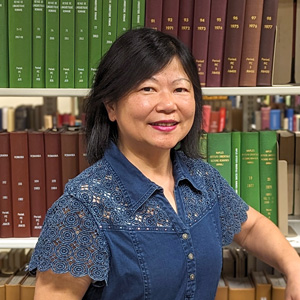 Photo of a woman with medium black hair and a blue shirt standing in front of a stack of books.