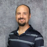 Man with very short hair and goatee wearing a dark blue striped shirt stading in front of a gray textured background