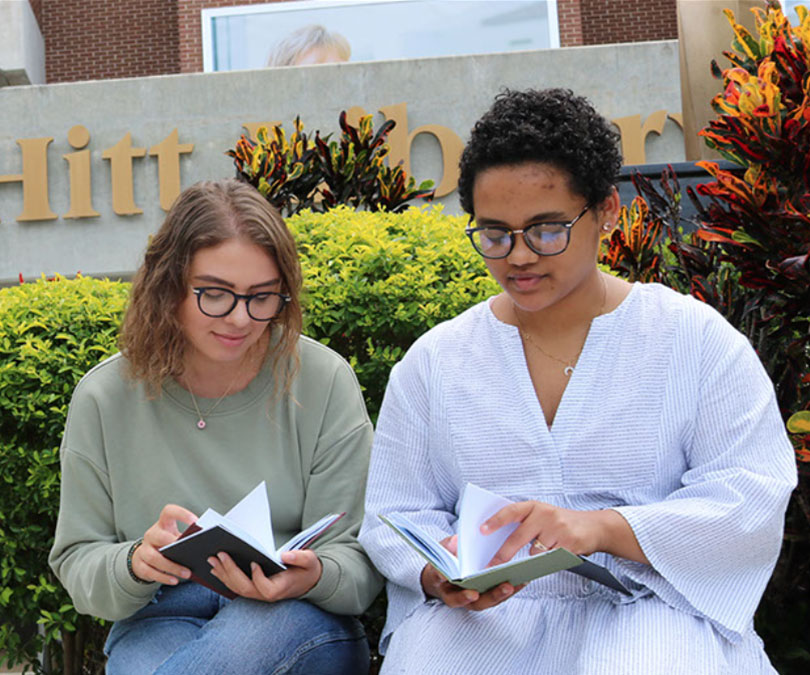 Two students looking at notebooks sitting in front of the John C. Hitt Library