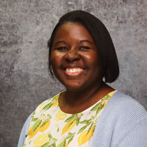 Friendly woman with medium length dark hair smiling warmly against a textured gray backdrop in a white, yellow, and green lemon pattern top and light blue sweater.