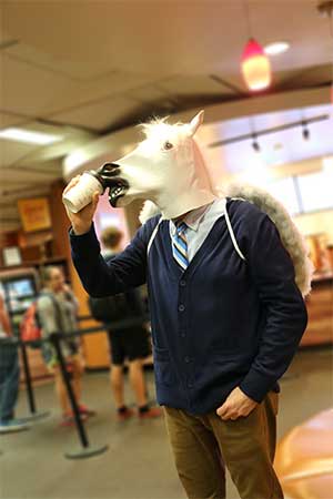 Photograph of a suave gentleprofessor wearing a white horse head mask while drinking a coffee in the Infusion Cafe of the Hitt Library