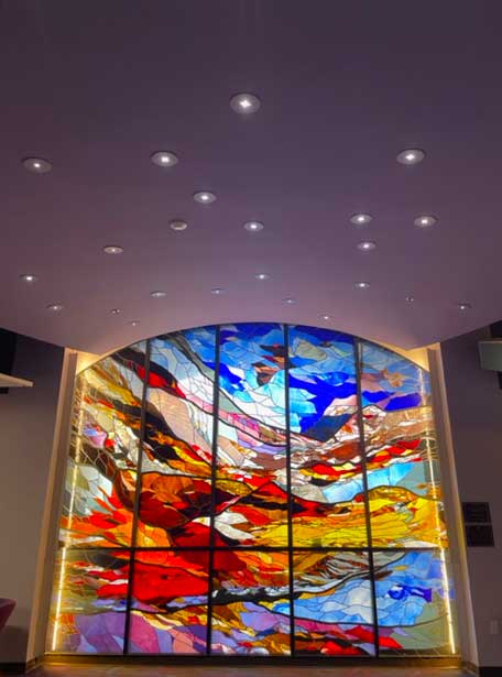 Photo of the colorful stained glass window titled Genesis on the third floor of the John C Hitt Library underneath a blue cieling with circular lights in the shape of the constellation of Pegsus.
