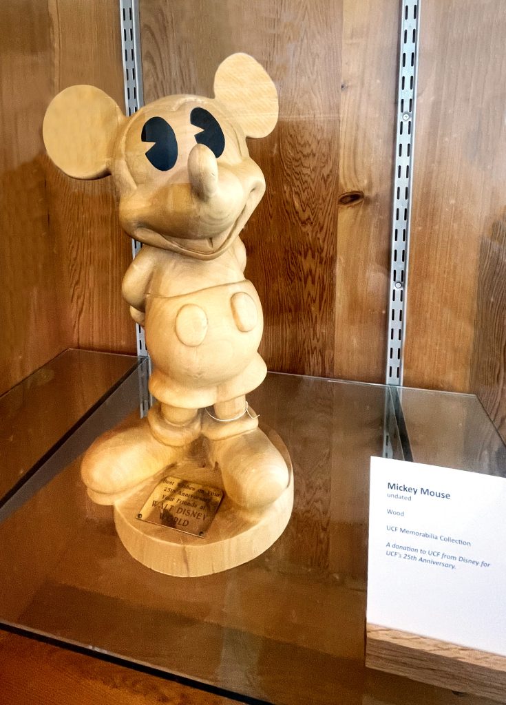 Photo of a wooden Mickey Mouse sculpture on a glass shelf in a wooden bookcase.