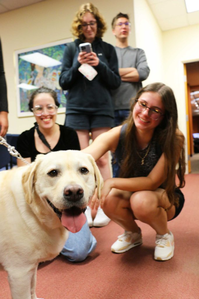 Photo of a yellow lab named Chipper with two students petting him