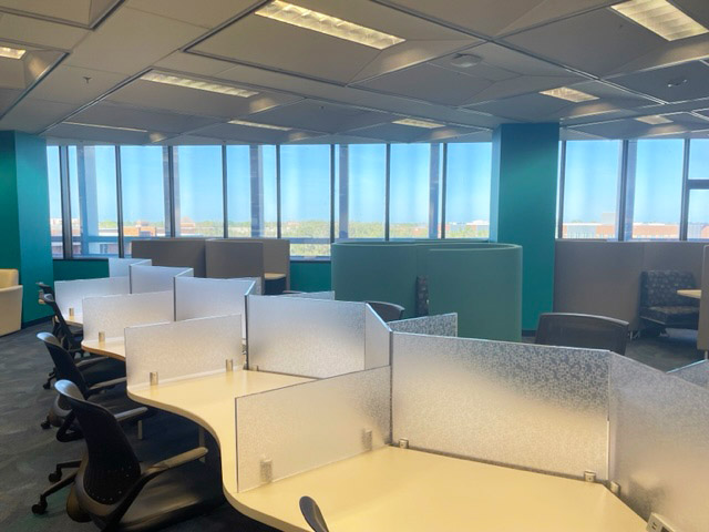 Photo of white tables with glass partitions against a backdrop of a long glass window on the 5th floor of the John C Hitt Library.