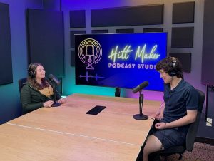 Two students with headphones and microphones recording a podcast with a large monitor behind them