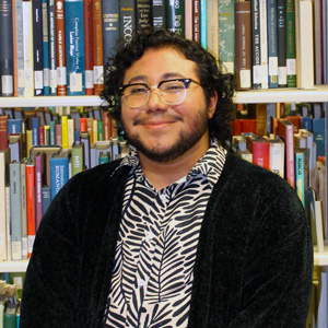 Photo of a man wearing a black and white striped shirt with a black sweater and glasses. Standing in front of rows of library books.