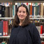 Woman with shoulder length, dark brown, hair in a blue blouse standing in front of a stack of books.