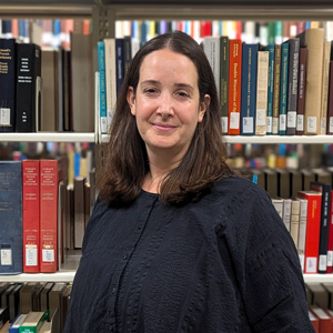 Woman with shoulder length, dark brown, hair in a blue blouse standing in front of a stack of books.