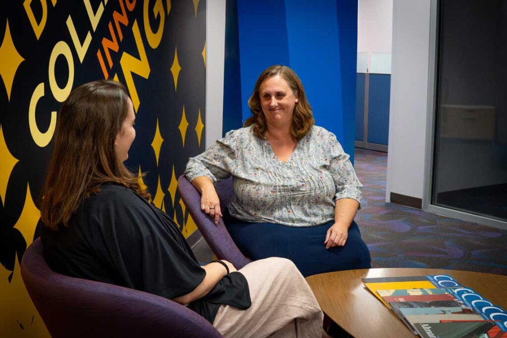 Two women engage in a thoughtful discussion in a modern office setting with colorful wall art during a sunny afternoon