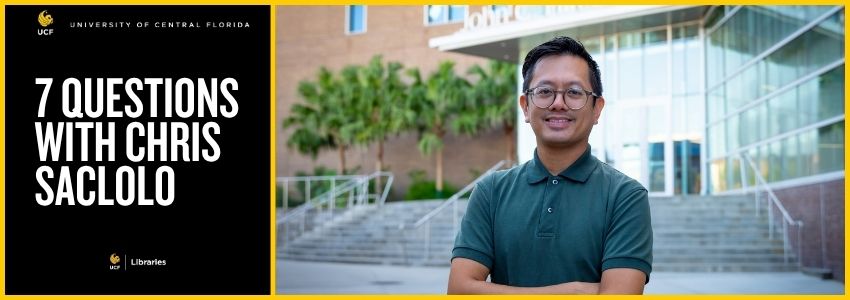 Man stands with arms crossed in front of Library entrance. He has glasses, dark hair and a green shirt.