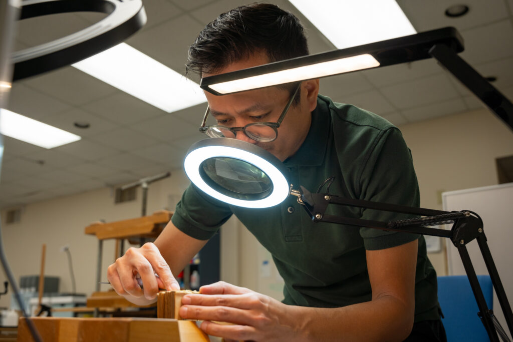 Man carefully mends a book binding in a workshop. He has glasses on and is using a magnifying glass with a light on it.