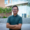 Man stands with arms crossed in front of Library entrance. He has glasses, dark hair and a green shirt.