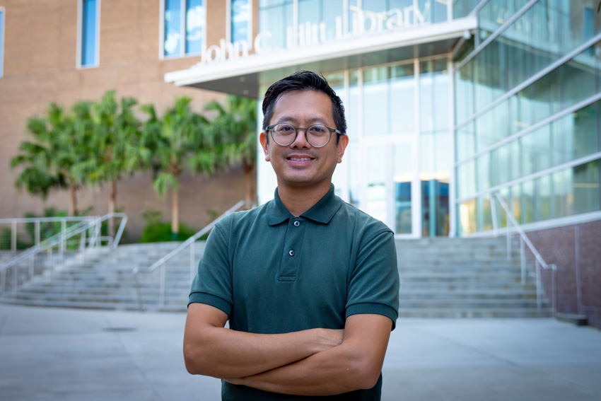 Man stands with arms crossed in front of Library entrance. He has glasses, dark hair and a green shirt.