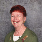 Friendly woman with short red hair smiling warmly against a textured gray backdrop in an olive green and white top