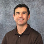 Friendly man with short dark hair smiling warmly against a textured gray backdrop in a dark brown polo shirt