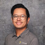 Friendly man with short black hair smiling warmly against a textured gray backdrop in a dark gray polo shirt.