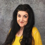 Friendly woman with long dark hair smiling warmly against a textured gray backdrop in a bright yellow top