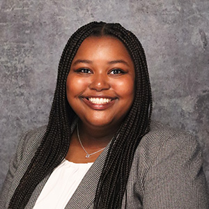 Friendly woman with long dark hair smiling warmly against a textured gray backdrop in grey and white pinstriped blazer and white blouse.