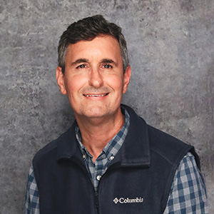 Friendly man with short dark hair smiling warmly against a textured gray backdrop in a blue checkered button down shirt with a dark blue sweater vest