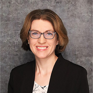 Friendly woman with medium length brown hair smiling warmly against a textured gray backdrop in a dark blue blazer and white blouse.