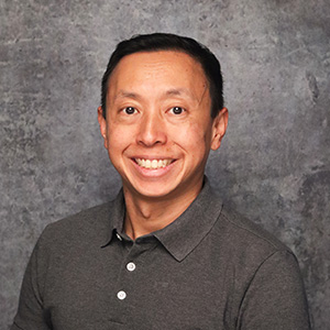 Friendly man with short black hair smiling warmly against a textured gray backdrop in a dark gray polo shirt.