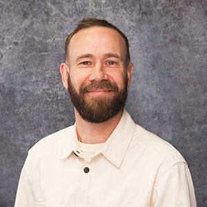 Friendly man with short brown hair smiling warmly against a textured gray backdrop in a beige collared shirt