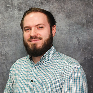 Friendly man with medium length brown hair and a full beard smiling slightly against a textured gray backdrop in a white and blue hatch striped button up collared shirt.