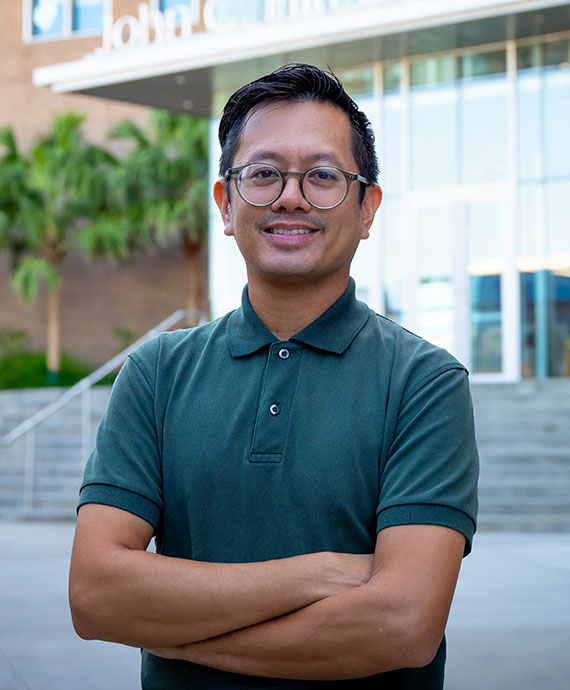 Chris Saclolo. A man with medium length black hair, round glasses, and a warm smile wearing a dark green polo shirt standing in front of the John C. Hitt Library entrance.