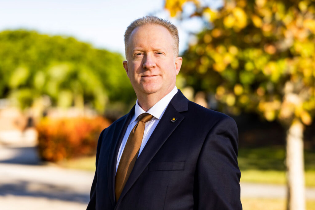 Man with short blond hair smiling slightly wearing a black suite and dark gold tie standing outside.