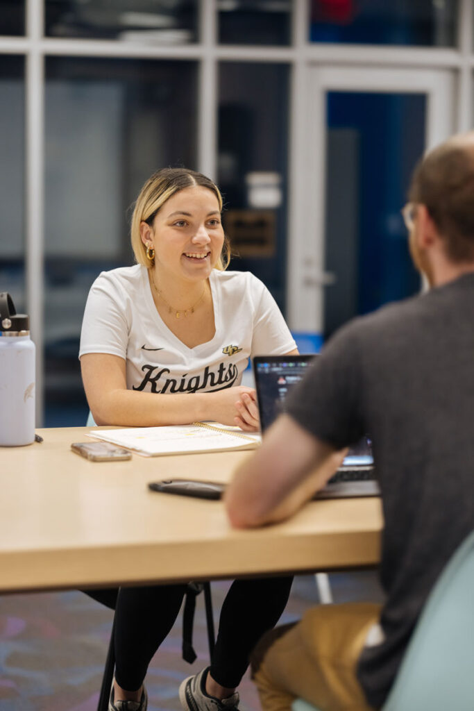 a female student with blond hair and a warm smile wearing a white UCF Knights t-shirt facing a male student with short brown hair and glasses and a dark gray t-shirt studying a table.