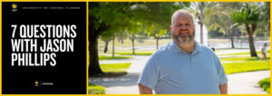 A man with a blue shirt smiling outside with a fountain behind him.