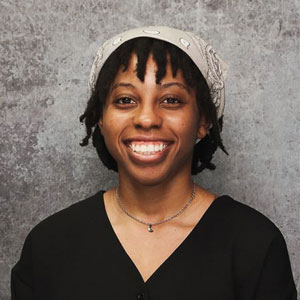 Friendly woman with medium length black hair smiling warmly against a textured gray backdrop in a black top.