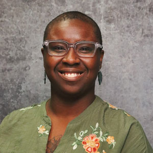Friendly person with very short dark hair smiling warmly against a textured gray backdrop in a green top with embroidered flowers.