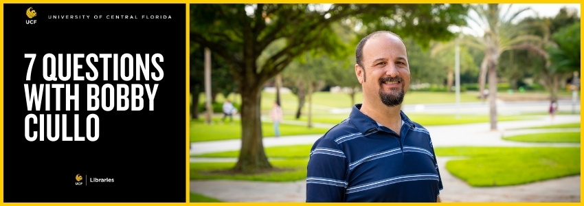 Man with striped shirt standing on a college campus with trees and a pond in the background.