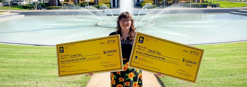 Woman standing with two large oversized checks in her hands in front of a fountain.