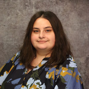 Friendly woman with long dark hair smiling against a textured gray backdrop in a dark blue blouse with light blue and yellow flower print.