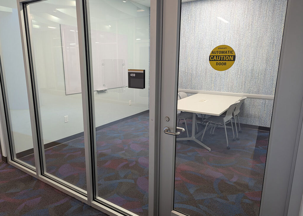 Study room with a rectangular table and four chairs on caster wheels with whiteboard mounted on the wall. Has a motion activated automatic door opener.