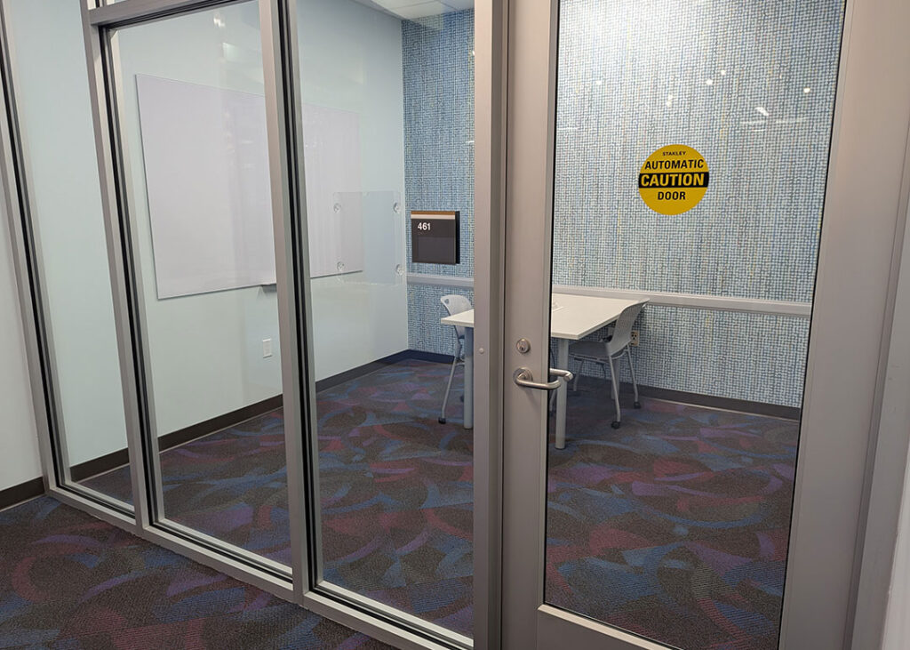 Study room with a rectangular table and two chairs on caster wheels with whiteboard mounted on the wall. Has a motion activated automatic door opener.