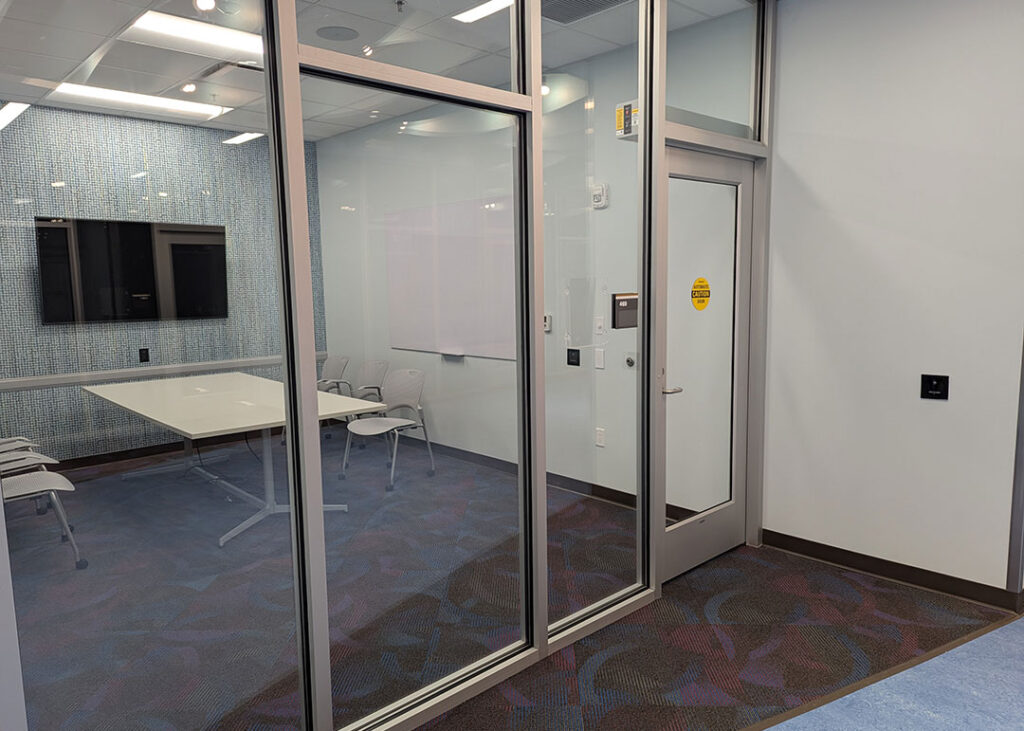 Study room with a rectangular table and sixchairs on caster wheels with whiteboard and monitor mounted on the walls. Has a motion activated automatic door opener.