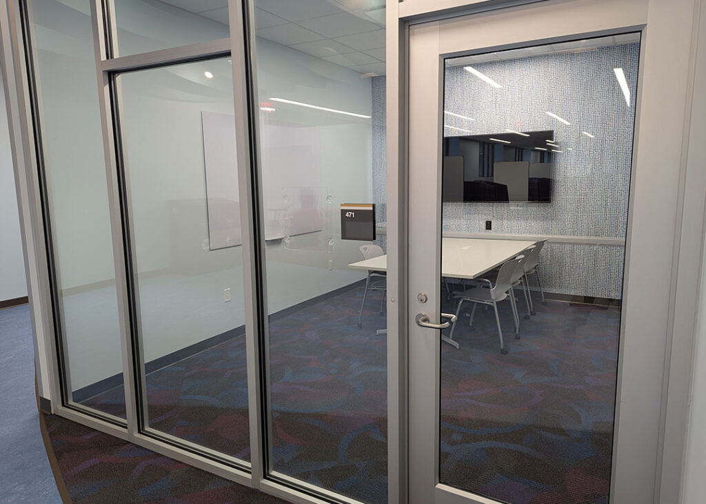 Study room with a rectangular table and sixchairs on caster wheels with whiteboard and monitor mounted on the walls. Has a motion activated automatic door opener.