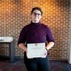 Girl standing in front of a brick wall with glasses on holding a certificate in front of her.