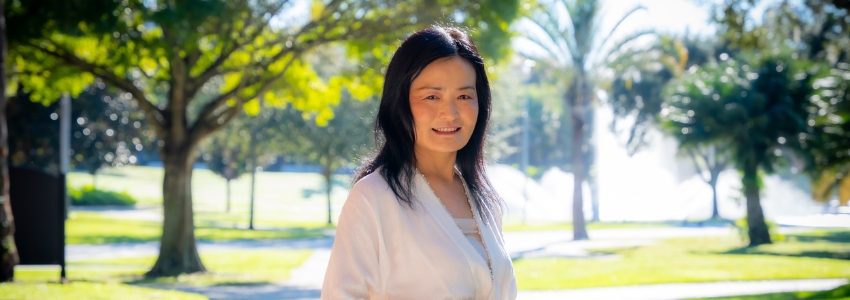 Woman with black hair and white shirt standing on college campus surrounded by trees.