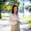 Woman with black hair and white shirt standing on college campus surrounded by trees.