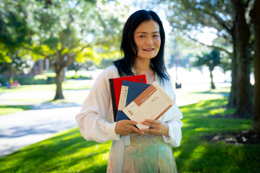 Woman with black hair and white shirt holding three books and standing on college campus surrounded by trees.