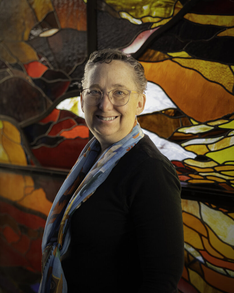 Woman with short hair and glasses stands in front of an abstract stained glass window with reds and yellows.