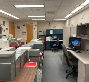 An office space with tables, archival boxes, computers, and filing cabinets under ceiling lights.