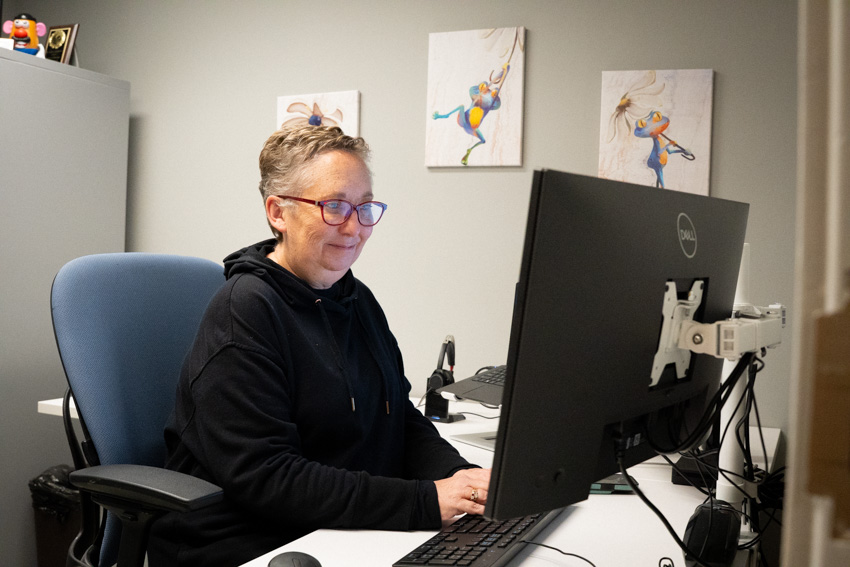 Woman in black sweatshirt with glasses and short hair sits at a desk working on a computer.